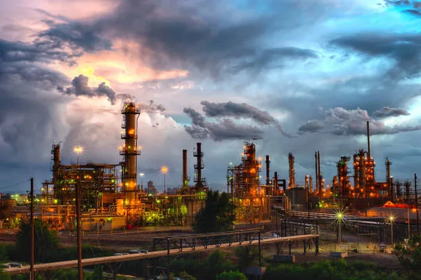 Illuminated refinery towers and pipework under a dramatic evening sky